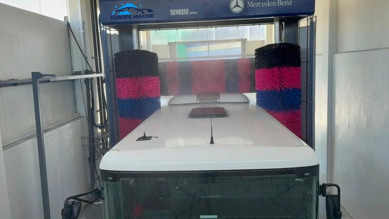 The roof of a white bus entering an indoor wash gantry with red, blue and black brushes.