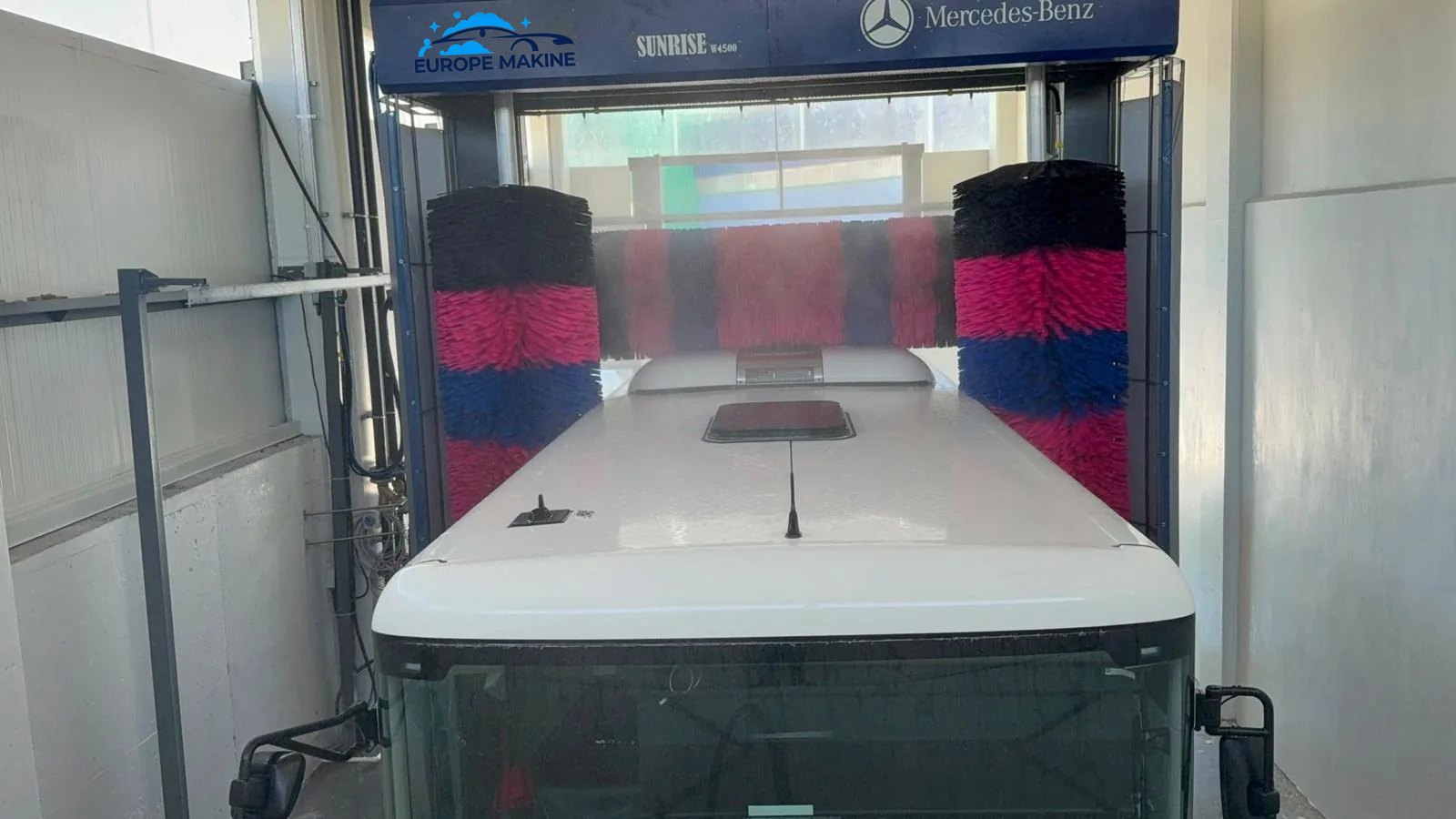 The roof of a white bus entering an indoor wash gantry with red, blue and black brushes.